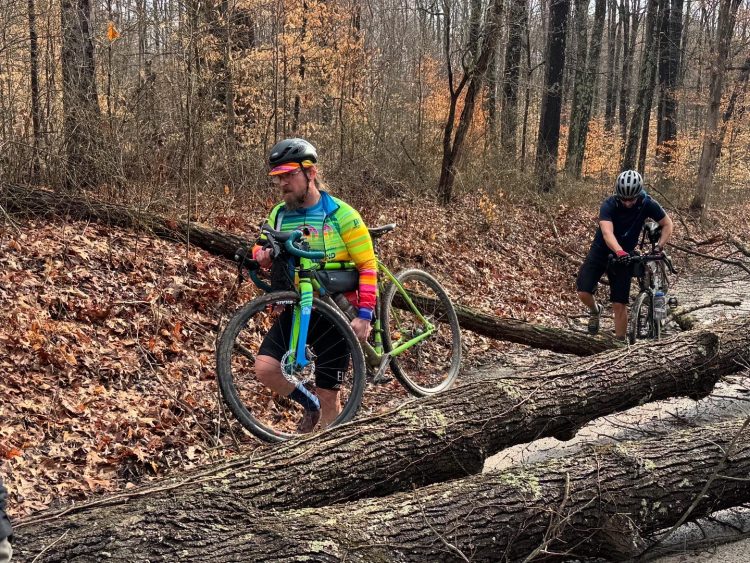 2 riders lifting cyclocross bikes over multiple trees covering the trail